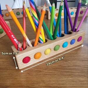 Puede incluir: Un portal&aacute;pices rectangular de madera con recortes circulares que contienen bolas de fieltro de colores y varios l&aacute;pices de colores. El soporte mide 24 cm de largo y 5 cm de alto, con un acabado de madera natural y detalles de fieltro vibrantes.