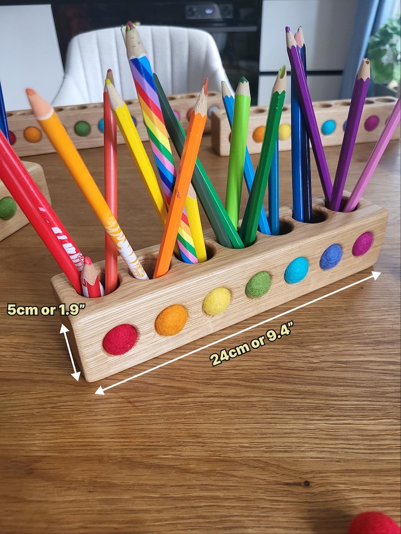 Puede incluir: Un portal&aacute;pices rectangular de madera con recortes circulares que contienen bolas de fieltro de colores y varios l&aacute;pices de colores. El soporte mide 24 cm de largo y 5 cm de alto, con un acabado de madera natural y detalles de fieltro vibrantes.