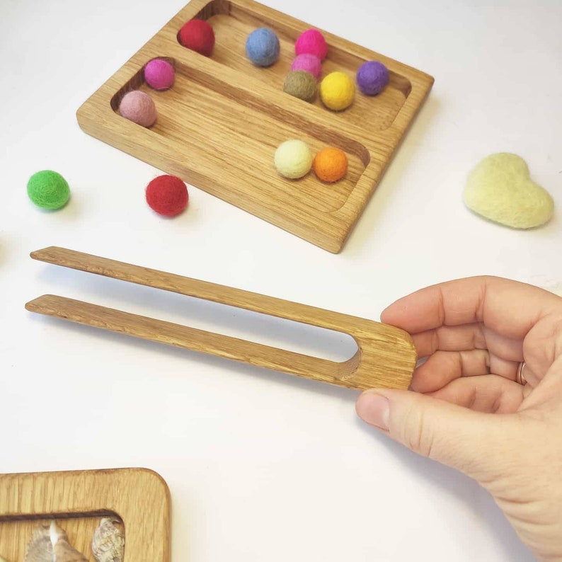 Puede incluir: Bandeja de madera con dos compartimentos llenos de bolas de fieltro de colores. Se muestra un par de pinzas de madera en primer plano.