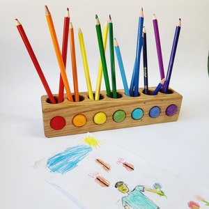 Puede incluir: Un portal&aacute;pices rectangular de madera con doce orificios circulares, cada uno con un l&aacute;piz de color. La parte frontal del soporte presenta seis incrustaciones circulares de fieltro en rojo, naranja, amarillo, verde, azul y morado. Un dibujo infantil se encuentra delante.