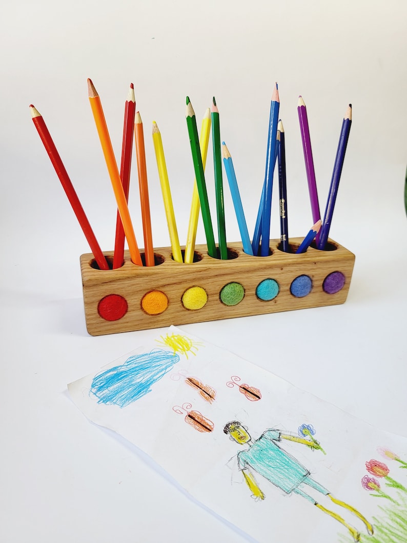 Puede incluir: Un portal&aacute;pices rectangular de madera con doce orificios circulares, cada uno con un l&aacute;piz de color. La parte frontal del soporte presenta seis incrustaciones circulares de fieltro en rojo, naranja, amarillo, verde, azul y morado. Un dibujo infantil se encuentra delante.