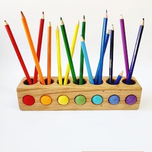 Puede incluir: Un portal&aacute;pices rectangular de madera clara con siete orificios circulares en la parte superior y siete bolas de lana fieltrada a juego en rojo, naranja, amarillo, verde, azul claro, azul y morado en el frente. El texto dice: pencils not included.