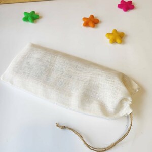 May include: A white linen drawstring bag with a brown cord. The bag is lying on a white surface with four felt stars in the background: one green, one orange, one yellow, and one pink.