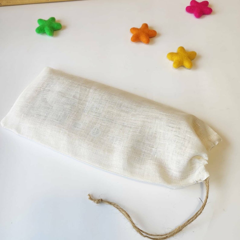 May include: A white linen drawstring bag with a brown cord. The bag is lying on a white surface with four felt stars in the background: one green, one orange, one yellow, and one pink.