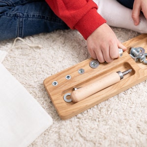 May include: A wooden tray with compartments for screws, nuts, and a screwdriver. The tray is made of light wood and has a natural finish. The screws and nuts are silver and the screwdriver has a wooden handle.