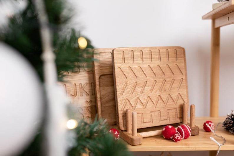May include: Wooden tracing boards with various patterns and letters, displayed on a wooden shelf. The boards are light brown and feature grooves for tracing. Red and white decorative balls are also on the shelf, along with a pinecone.