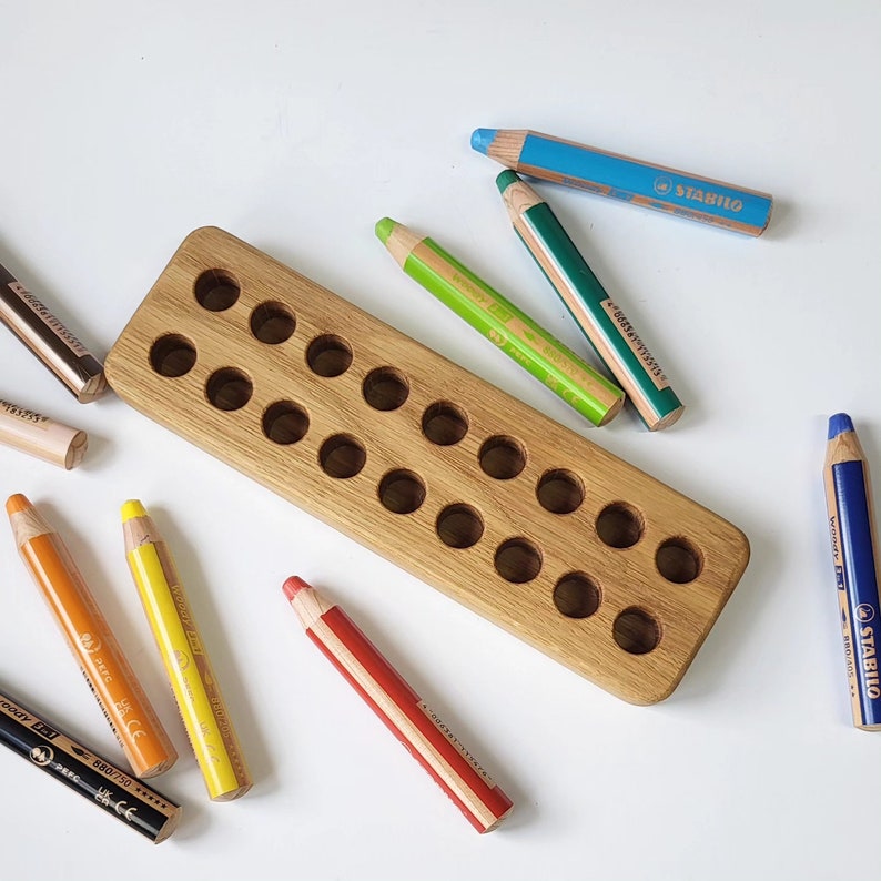 May include: A wooden pencil holder with multiple round holes, surrounded by colorful pencils. The pencils are various colors, including blue, green, yellow, orange, and red. Some pencils have the brand name "STABILO" printed on them.
