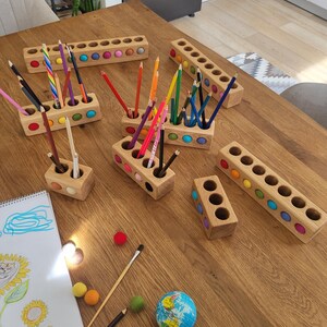 Puede incluir: Una colecci&oacute;n de portal&aacute;pices rectangulares de madera con aberturas circulares, cada uno con bolas de fieltro de colores incrustadas en los laterales. Los soportes contienen l&aacute;pices de colores y est&aacute;n dispuestos junto a un bloc de dibujo, bolas de fieltro sueltas y un peque&ntilde;o globo terr&aacute;queo.