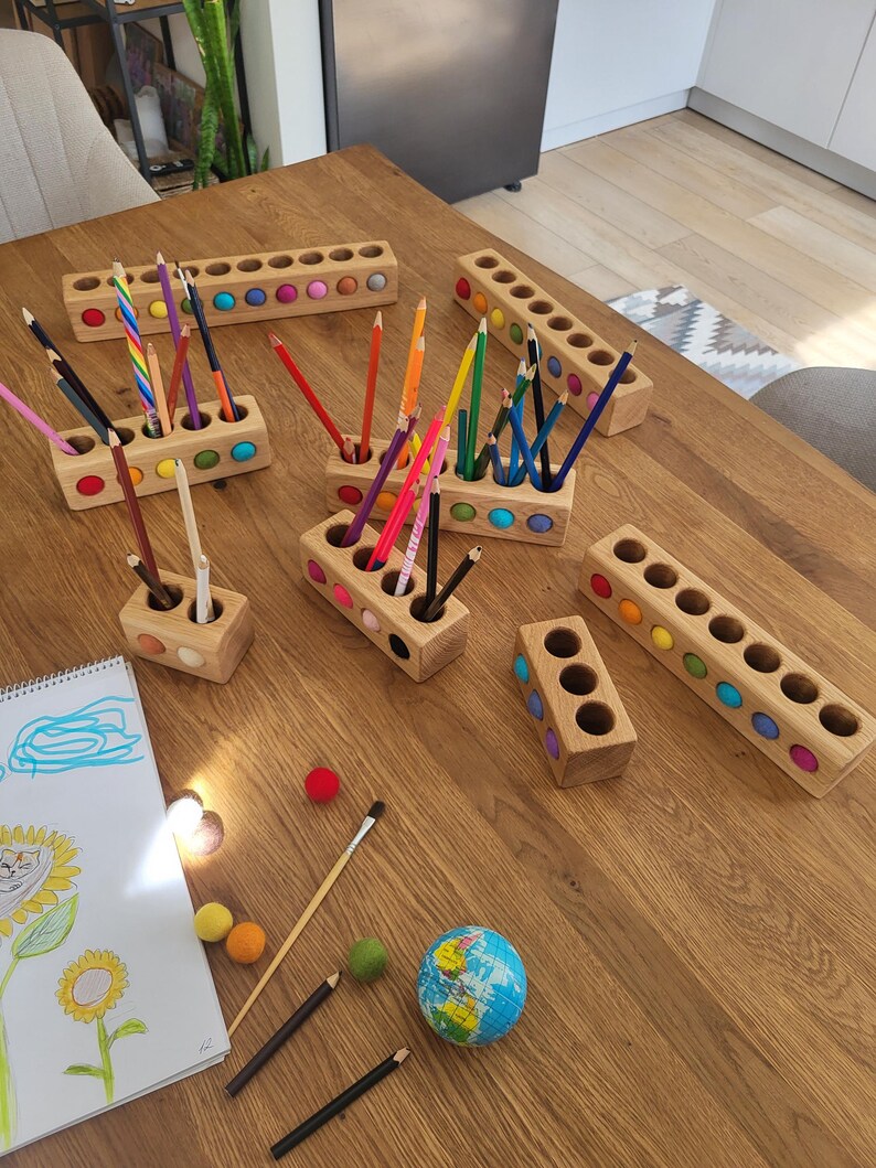 Puede incluir: Una colecci&oacute;n de portal&aacute;pices rectangulares de madera con aberturas circulares, cada uno con bolas de fieltro de colores incrustadas en los laterales. Los soportes contienen l&aacute;pices de colores y est&aacute;n dispuestos junto a un bloc de dibujo, bolas de fieltro sueltas y un peque&ntilde;o globo terr&aacute;queo.