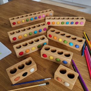 Puede incluir: Una colecci&oacute;n de bloques rectangulares de madera con orificios circulares, cada uno con una peque&ntilde;a bola de fieltro de varios colores. Los bloques var&iacute;an en longitud, con filas de dos a diez orificios, algunos de los cuales est&aacute;n vac&iacute;os.