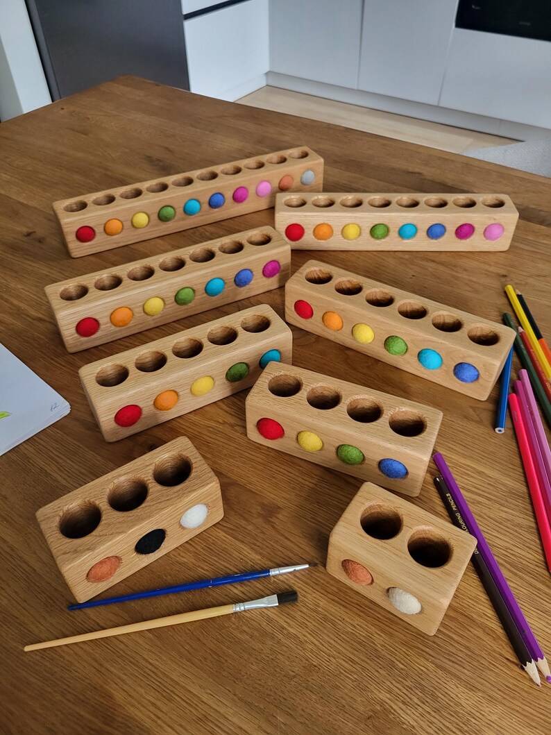 Puede incluir: Una colecci&oacute;n de bloques rectangulares de madera con orificios circulares, cada uno con una peque&ntilde;a bola de fieltro de varios colores. Los bloques var&iacute;an en longitud, con filas de dos a diez orificios, algunos de los cuales est&aacute;n vac&iacute;os.