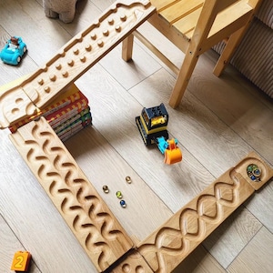 Op de afbeelding: Houten knikkerbaan met verschillende baanontwerpen, waaronder een zigzag en een reeks pinnen. Een blauwe speelgoedauto en een gele en zwarte speelgoedgraafmachine liggen op de grond. Een knuffelschaap en een houten stoel staan ook in de afbeelding.