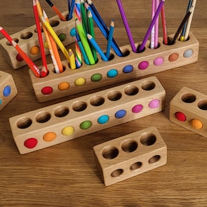 Könnte beinhalten: Stifthalter aus Holz mit farbigen Filzakzenten. Die Halter sind rechteckig mit mehreren kreisförmigen Öffnungen für Bleistifte und Stifte. Jeder Halter hat eine Reihe von Filzkugeln in verschiedenen Farben, darunter Rot, Orange, Gelb, Grün, Blau und Lila.
