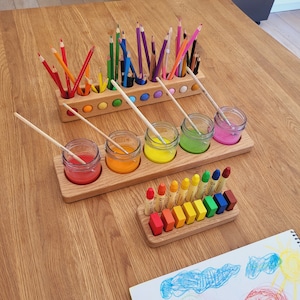 Puede incluir: Juego de materiales de arte de madera con lápices de colores, botes de pintura y ceras. Los lápices están ordenados por color en un soporte de madera. Los botes de pintura contienen pintura roja, naranja, amarilla, verde y rosa. Las ceras también están ordenadas por color.