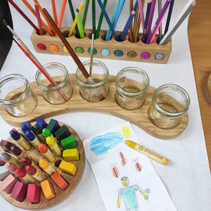 May include: A wooden pencil holder with colourful felt inserts, a curved wooden stand holding five glass jars, and a circular wooden block containing various wax crayons. A yellow Stockmar crayon and two coloured pencils lie on a drawing.