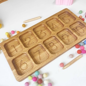 Könnte beinhalten: Holztablett mit zehn Fächern, die jeweils eine Zahl von 1 bis 10 enthalten. Das Tablett ist aus hellbraunem Holz gefertigt und hat eine natürliche Oberfläche. Es gibt bunte Kugeln, die um das Tablett herum verstreut sind.