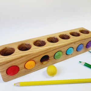 Puede incluir: Un bloque rectangular de madera con ocho orificios circulares perforados en la parte superior y frontal. El bloque presenta bolas de fieltro en colores rojo, naranja, amarillo, verde, azul y morado, con un l&aacute;piz amarillo y uno verde cerca.