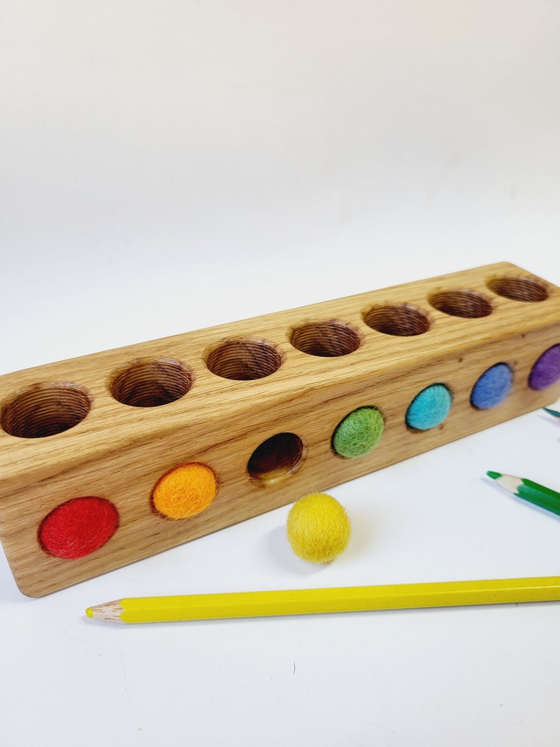 Puede incluir: Un bloque rectangular de madera con ocho orificios circulares perforados en la parte superior y frontal. El bloque presenta bolas de fieltro en colores rojo, naranja, amarillo, verde, azul y morado, con un l&aacute;piz amarillo y uno verde cerca.