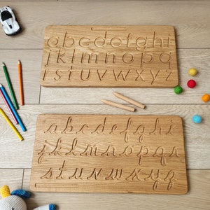 May include: Two wooden alphabet tracing boards with lowercase letters carved into the surface. One board features print letters, the other cursive. The boards are light brown and sit on a wooden floor, with pencils and toys nearby.
