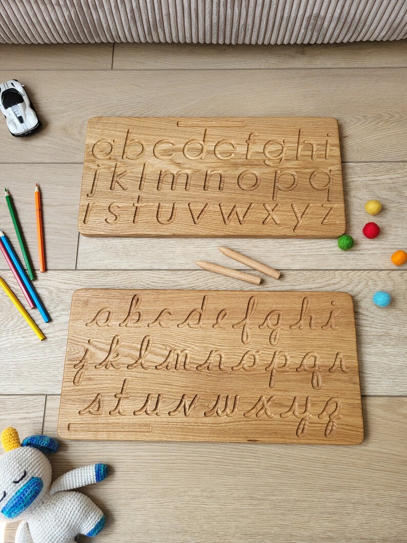 May include: Two wooden alphabet tracing boards with lowercase letters carved into the surface. One board features print letters, the other cursive. The boards are light brown and sit on a wooden floor, with pencils and toys nearby.