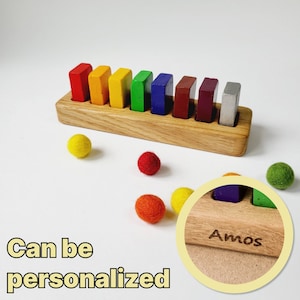 May include: A wooden rainbow stacking toy with colorful blocks and a personalized inscription that reads "Amos". The toy is accompanied by several small felt balls in various colors.