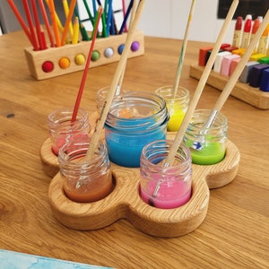Peut inclure: Un ensemble de fournitures d'art en bois avec six petits pots en verre remplis de peinture colorée et de pinceaux. Un porte-crayons en bois avec des crayons de couleur arc-en-ciel est également visible. L'ensemble est sur une table en bois.
