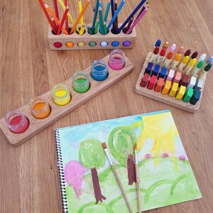 Puede incluir: Un portal&aacute;pices de madera con l&aacute;pices de colores, un soporte de madera con cinco frascos de vidrio con pintura, un bloque de madera con ceras rectangulares y un cuaderno de espiral con una acuarela y dos pinceles.