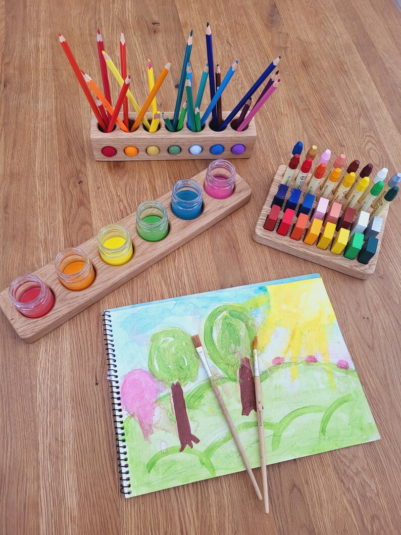 Puede incluir: Un portal&aacute;pices de madera con l&aacute;pices de colores, un soporte de madera con cinco frascos de vidrio con pintura, un bloque de madera con ceras rectangulares y un cuaderno de espiral con una acuarela y dos pinceles.