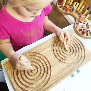 May include: A wooden board with two circular grooves for tracing with a wooden stick. The board is light brown and the grooves are dark brown. A child is tracing the grooves with the stick.