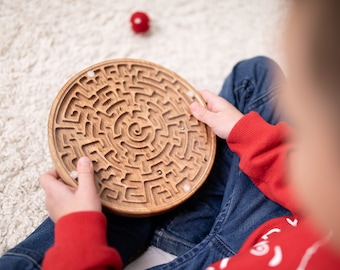 Puede incluir: Un niño juega con un juego de laberinto circular de madera. El laberinto tiene un patrón en espiral y está hecho de madera marrón claro. El niño lleva un suéter rojo y pantalones vaqueros azules.