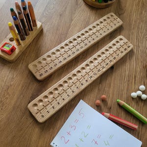 Może przedstawiać: Drewniane narzędzia edukacyjne do matematyki, w tym dwie osie liczbowe z otworami i oznaczeniami od -20 do 20. Kolorowe ołówki i notatnik z zadaniami matematycznymi również obecne, wszystko na drewnianej powierzchni.