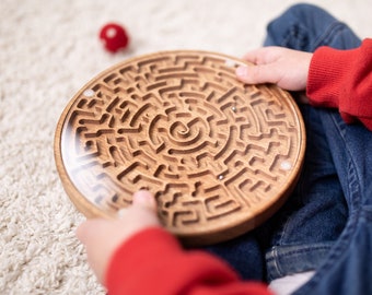 Puede incluir: Un juego de laberinto de madera con una cubierta de plástico transparente. El laberinto es intrincado y presenta un diseño en espiral en el centro. El juego está siendo sostenido por una persona que lleva ropa roja y azul.