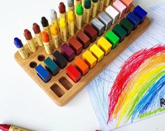May include: Wooden crayon organiser with a variety of colourful crayons and crayon blocks. The organiser holds crayons of different colours, including red, orange, yellow, green, blue, and purple. A drawing of a rainbow is on the right. The text "crayons not included" is at the bottom.