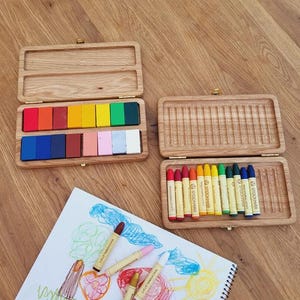 May include: Two wooden crayon boxes with colorful crayons. One box is open, revealing a variety of crayons in shades of red, orange, yellow, green, blue, and pink. The other box is open, showing a set of crayons. A drawing with crayons is on a white paper. The text "crayons not included" is visible.