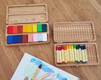 Puede incluir: Dos cajas de madera para lápices de colores con lápices de colores. Una caja está abierta, revelando una variedad de lápices de colores en tonos de rojo, naranja, amarillo, verde, azul y rosa. La otra caja está abierta, mostrando un juego de lápices de colores. Un dibujo con lápices de colores está sobre un papel blanco. El texto "crayons not included" es visible.