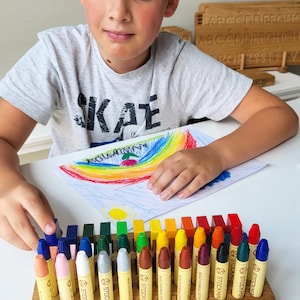 May include: A wooden box with 24 crayons arranged in rows. The crayons are labeled with the brand name "STOCKMAR" and the words "WACHSMALSTIFT". The crayons are in a variety of colors, including red, orange, yellow, green, blue, purple, brown, and black.