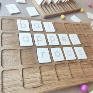 May include: Wooden educational toy featuring a light brown wooden tray with recessed squares. White rectangular tiles with engraved lowercase letters spell out "ball" and "appels". Additional letter tiles and small felt balls are scattered around the tray.