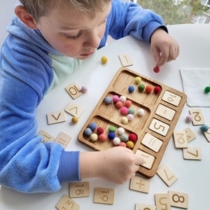 May include: A wooden educational math board with carved numbers and symbols, and a section for colorful felt balls. The board is surrounded by individual wooden number tiles and felt balls. A child is interacting with the educational toy.