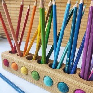Puede incluir: Un portal&aacute;pices rectangular de madera con una fila de orificios circulares que contienen l&aacute;pices de colores en tonos rojo, naranja, amarillo, verde, azul y morado. El frente del soporte presenta una fila de peque&ntilde;os insertos de lana fieltrada.