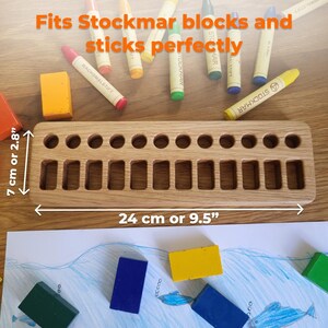 May include: A wooden crayon holder, 9.5 inches long and 2.8 inches wide, with 14 rectangular and 14 circular slots. The text "Fits Stockmar blocks and sticks perfectly" is visible. Several colorful crayons and blocks are scattered around the holder.