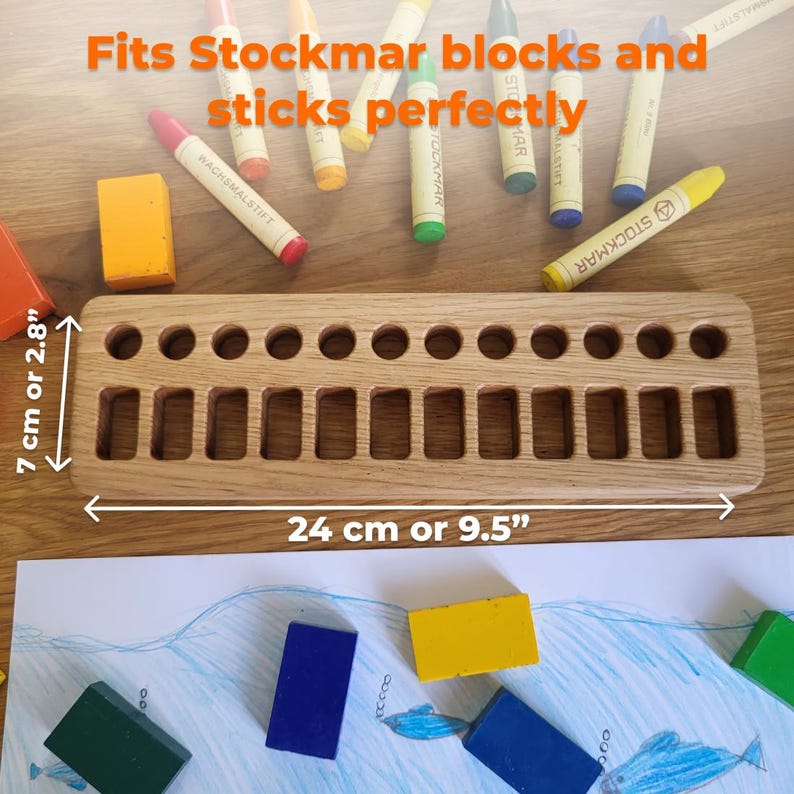 May include: A wooden crayon holder, 9.5 inches long and 2.8 inches wide, with 14 rectangular and 14 circular slots. The text "Fits Stockmar blocks and sticks perfectly" is visible. Several colorful crayons and blocks are scattered around the holder.