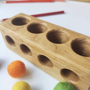 Puede incluir: Un bloque de madera rectangular de color marr&oacute;n claro con una serie de orificios circulares perforados en las superficies superior y frontal, acompa&ntilde;ado de varias bolas peque&ntilde;as de lana fieltrada en colores naranja, amarillo y verde.