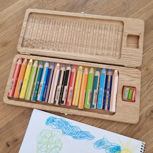 May include: Wooden pencil case with a hinged lid, containing a set of colorful pencils. A pencil sharpener is visible. A drawing with colorful crayon marks is in the foreground. The text "pencils not included" is at the bottom.