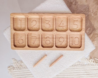 Puede incluir: Bandeja de madera con diez compartimentos numerados, cada uno con un número del 1 al 10. Dos palitos de madera se colocan sobre una superficie blanca junto a la bandeja.