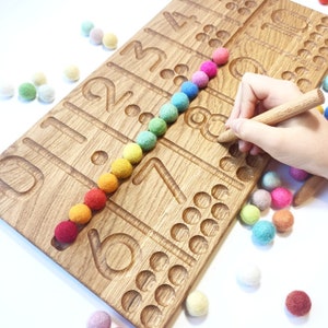 May include: Wooden number tracing board with carved numbers and corresponding dots. A row of colorful felt balls in rainbow colors is placed along the board. A wooden stylus is used to trace the number 8. Additional felt balls are scattered around the board.