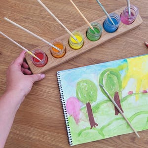 May include: A wooden paint palette with six small glass jars filled with colourful paint. Paintbrushes are placed in each jar. A watercolour painting of trees and a paintbrush are on the wooden surface.