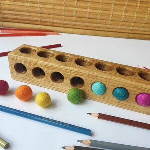 Puede incluir: Un bloque de madera rectangular de color marr&oacute;n claro con dos filas de orificios circulares. Varias bolas peque&ntilde;as de lana fieltrada de colores vibrantes, incluyendo rojo, naranja, amarillo, verde, azul y magenta, est&aacute;n cerca del bloque.