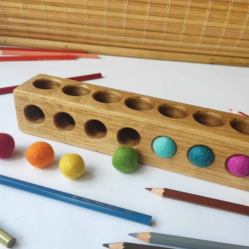 Puede incluir: Un bloque de madera rectangular de color marr&oacute;n claro con dos filas de orificios circulares. Varias bolas peque&ntilde;as de lana fieltrada de colores vibrantes, incluyendo rojo, naranja, amarillo, verde, azul y magenta, est&aacute;n cerca del bloque.