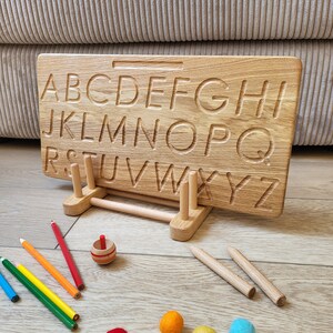 May include: A wooden alphabet tracing board with carved letters, supported by a wooden stand. Colorful pencils, wooden dowels, a spinning top, and felt balls in various colors are scattered around the board. The board is designed for educational play.
