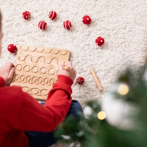 May include: A wooden tracing board with carved lines and circles, and a wooden stylus. Scattered around are red and white felt balls. The scene is set on a white rug, with a Christmas tree in the background.
